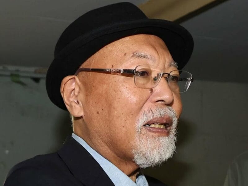 Close-up of an older man with a white beard wearing a black hat and glasses, speaking at an event.