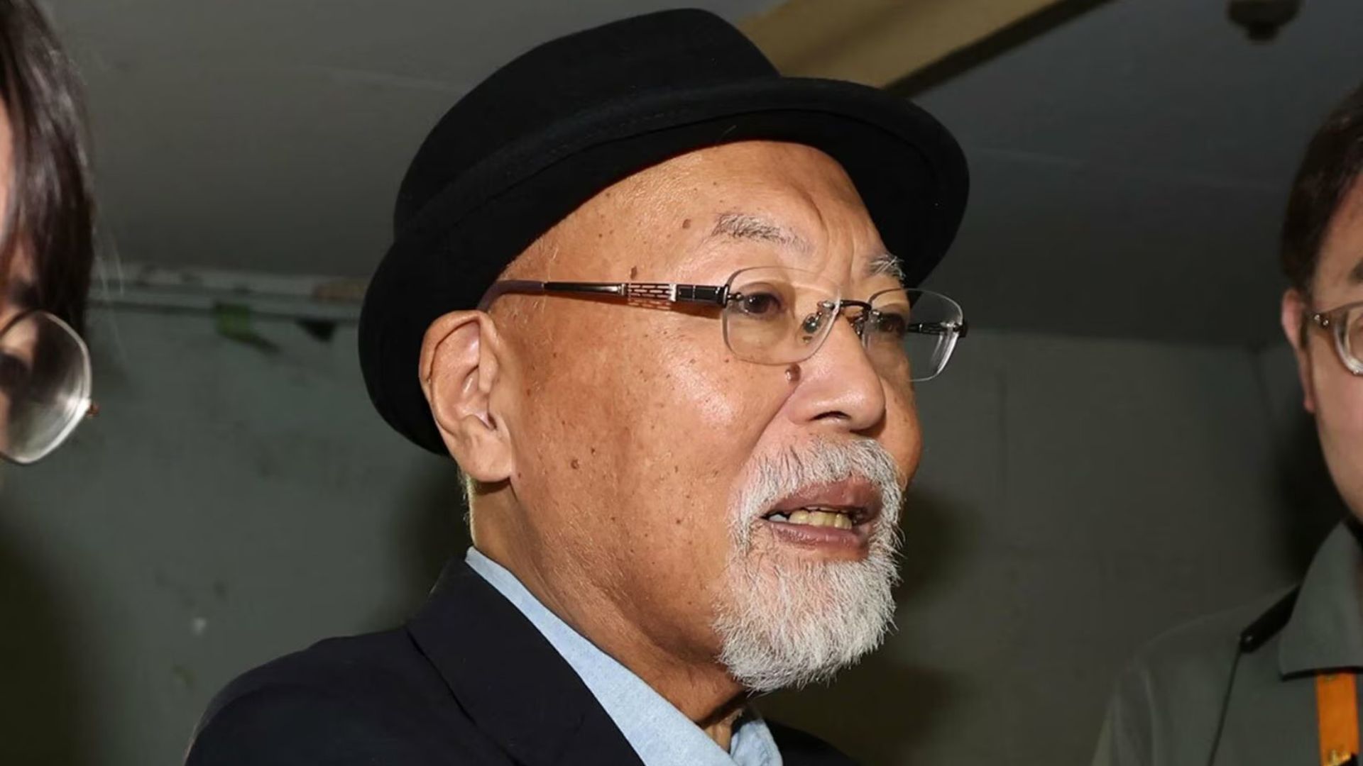 Close-up of an older man with a white beard wearing a black hat and glasses, speaking at an event.