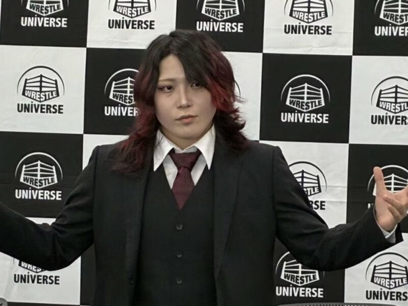 Person in a dark suit and maroon tie stands in front of a Wrestle Universe checkered backdrop, arms extended to the sides.