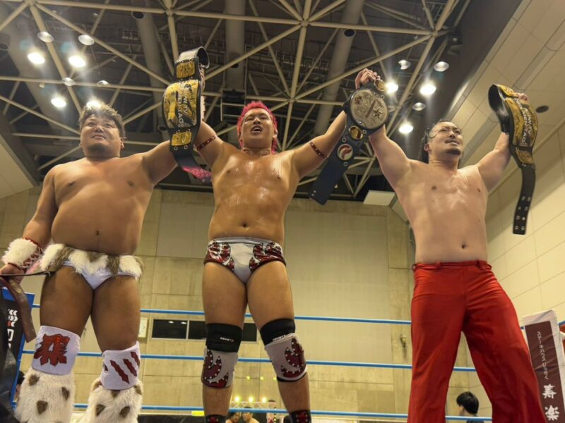 Three wrestlers stand in a ring, raising their championship belts in celebration after a victory inside a gym arena.