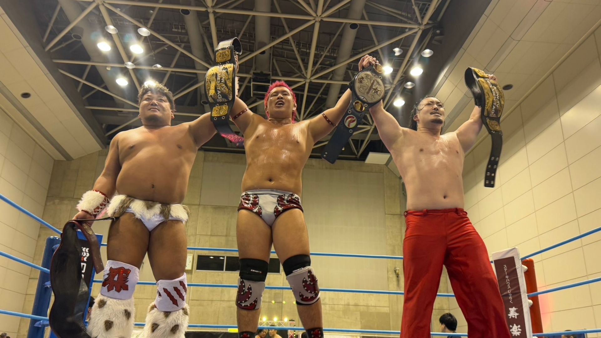 Three wrestlers stand in a ring, raising their championship belts in celebration after a victory inside a gym arena.