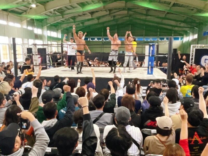 Wrestlers celebrate in the ring as a large cheering crowd raises fists behind them in a gym-style arena.