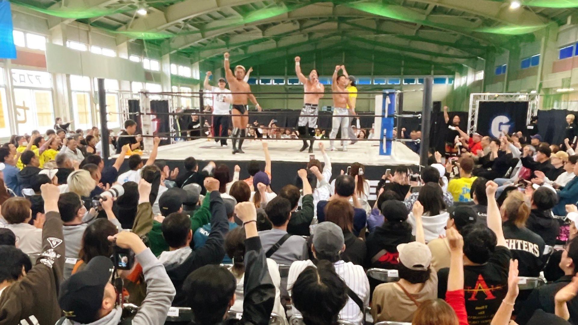 Wrestlers celebrate in the ring as a large cheering crowd raises fists behind them in a gym-style arena.