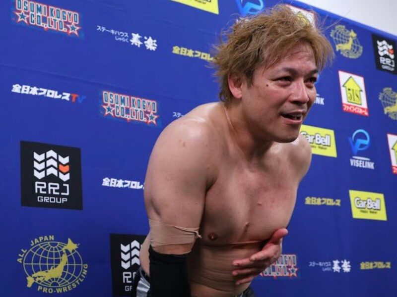 Shirtless pro wrestler leaning forward in front of a blue sponsor backdrop after a match.