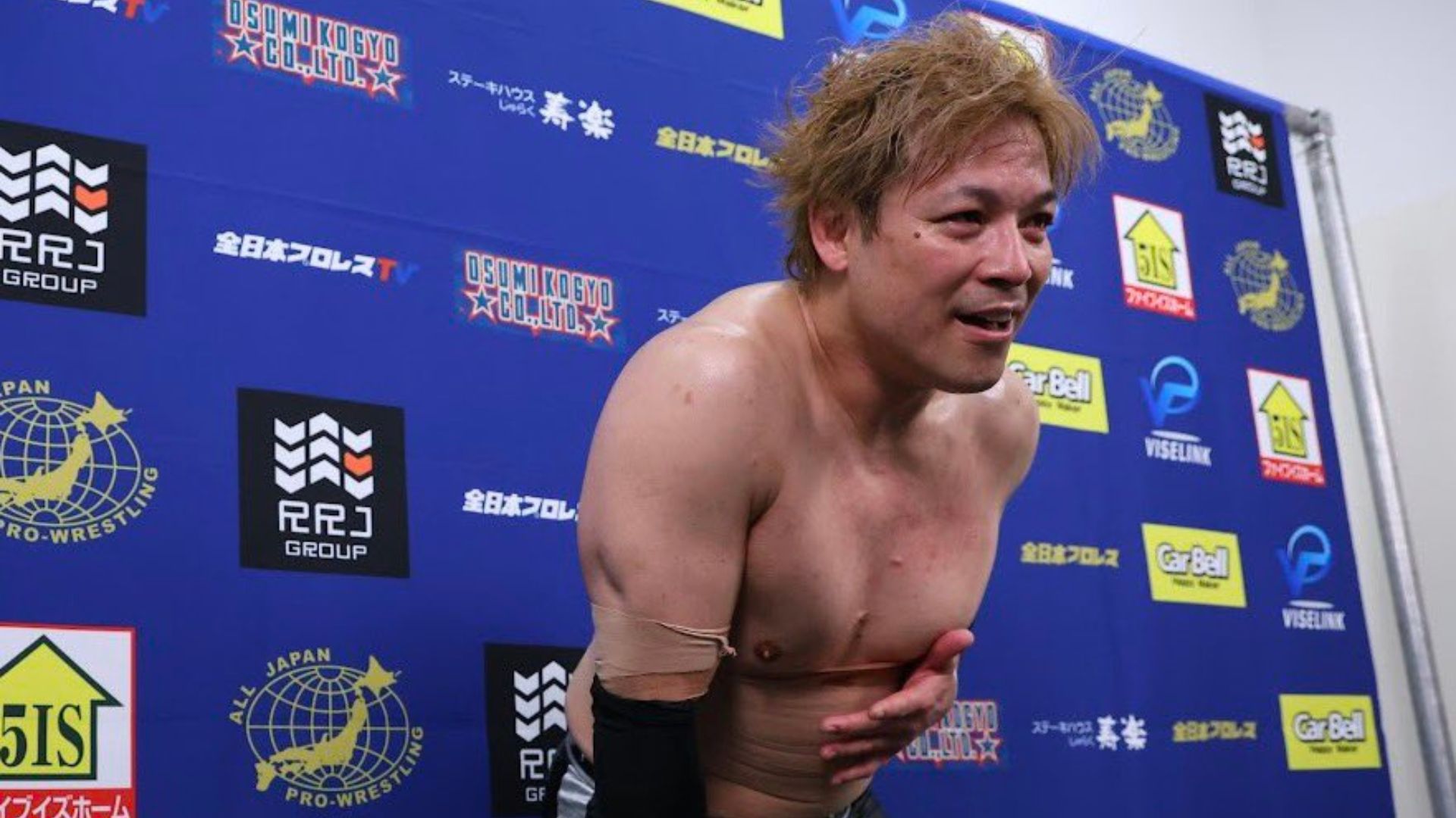 Shirtless pro wrestler leaning forward in front of a blue sponsor backdrop after a match.