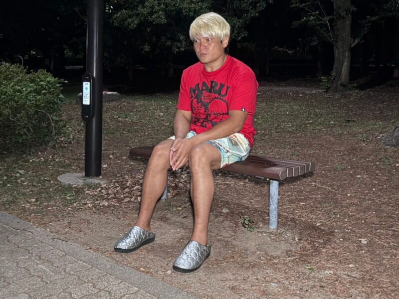 Man with a blonde wig sits on a park bench at night, wearing a red graphic t-shirt and light shorts.