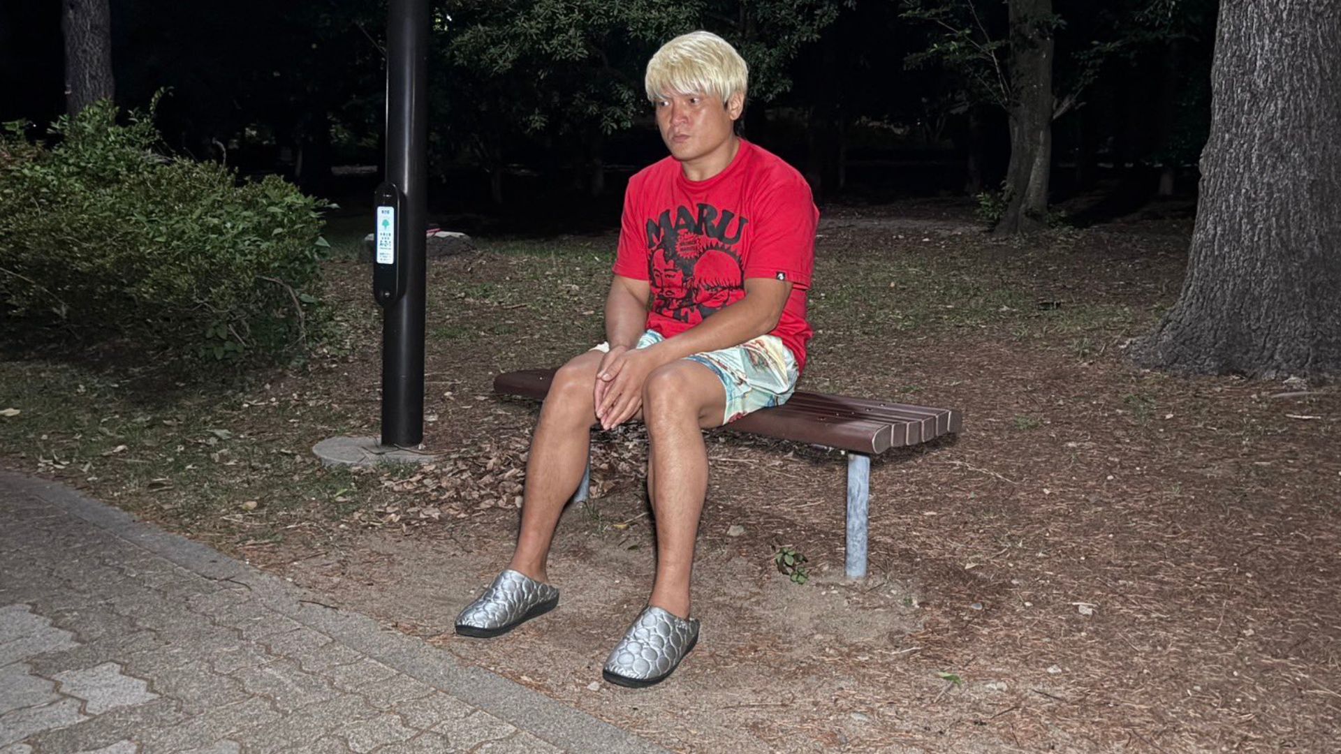 Man with a blonde wig sits on a park bench at night, wearing a red graphic t-shirt and light shorts.