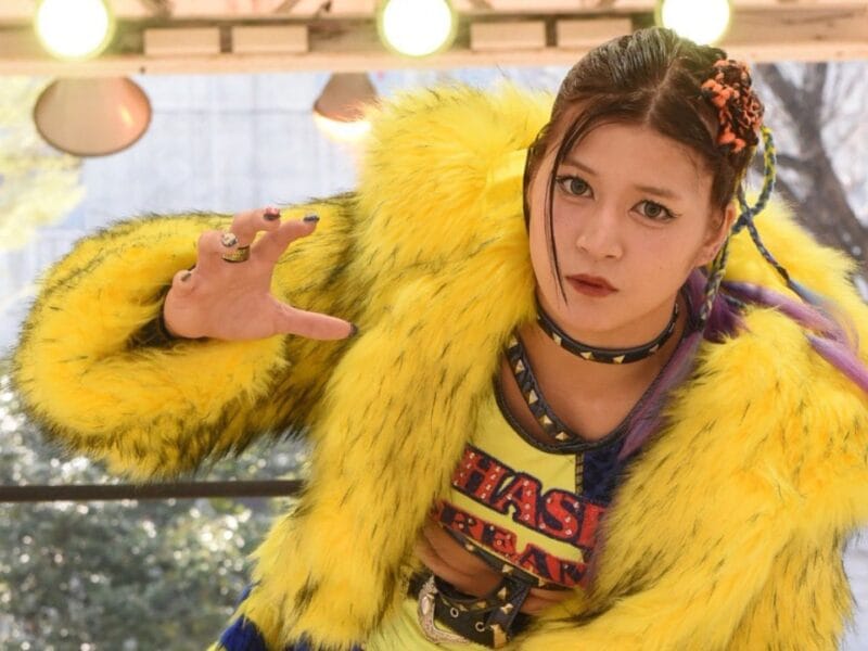 Person in a bright yellow fur coat and braided hair extends a hand toward the camera outdoors, posing dramatically.