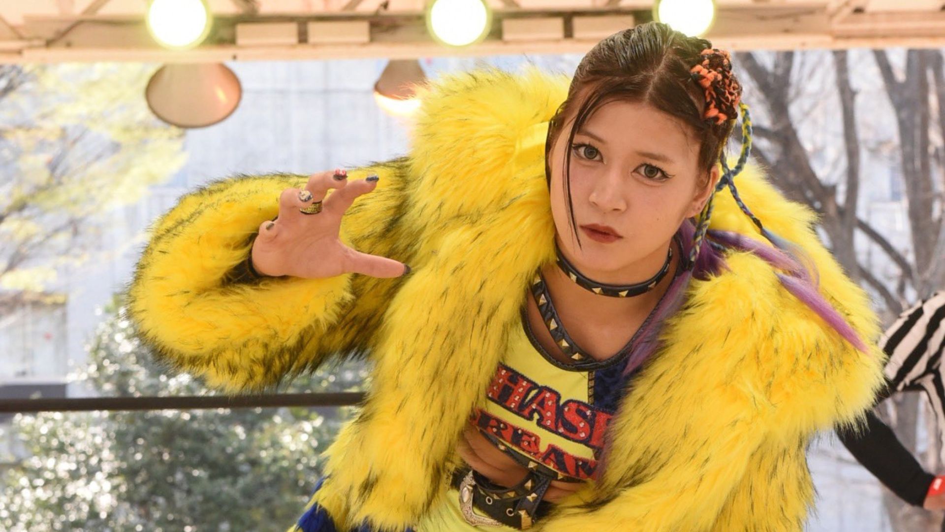 Person in a bright yellow fur coat and braided hair extends a hand toward the camera outdoors, posing dramatically.