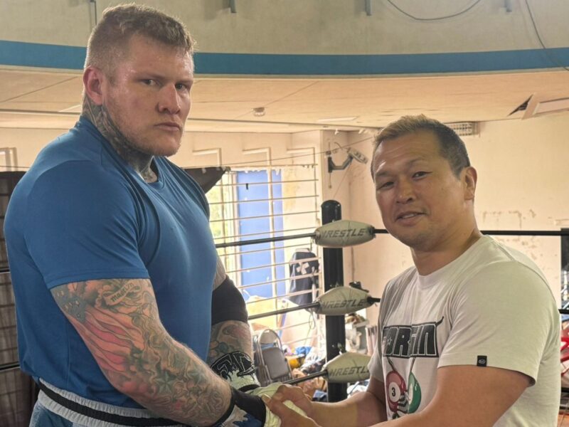 Two muscular men shake hands inside a boxing/wrestling gym, wearing athletic gear and tattoos visible on the left man's arms.