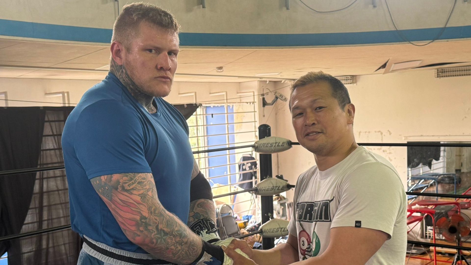 Two muscular men shake hands inside a boxing/wrestling gym, wearing athletic gear and tattoos visible on the left man's arms.
