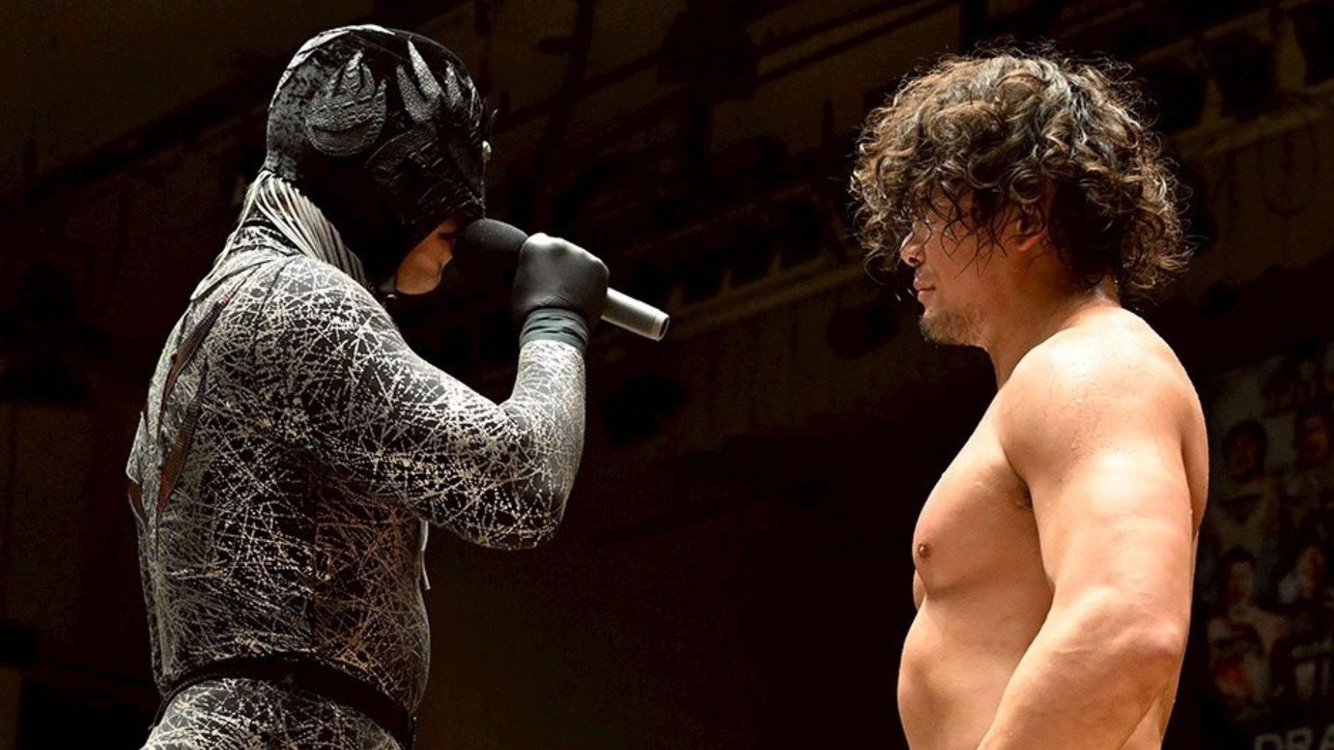 in a textured suit holds a microphone to a shirtless man facing him.