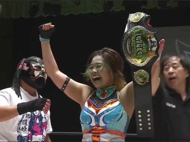 Female wrestler celebrates victory, raising a large championship belt above her head on stage.