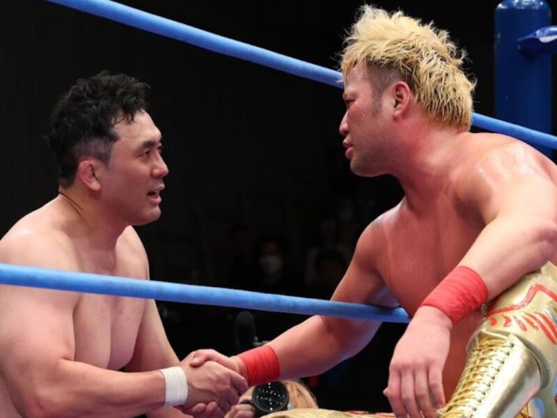 Two shirtless wrestlers shake hands in a wrestling ring after a match, with blue ropes framing them.