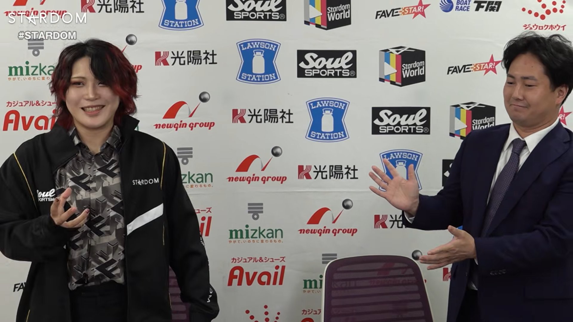 Two people pose in front of a wall covered with sponsor logos; left person smiles with red-dyed hair and a black jacket, right person in a suit gestures with hands downstage-right.