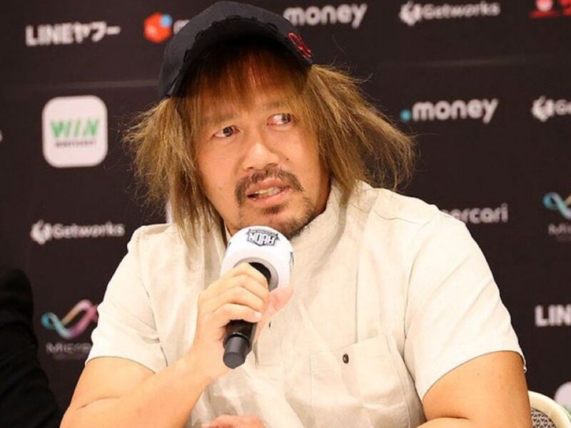 Man with shaggy hair and a cap speaking into a handheld microphone at a press conference backdrop full of sponsor logos.