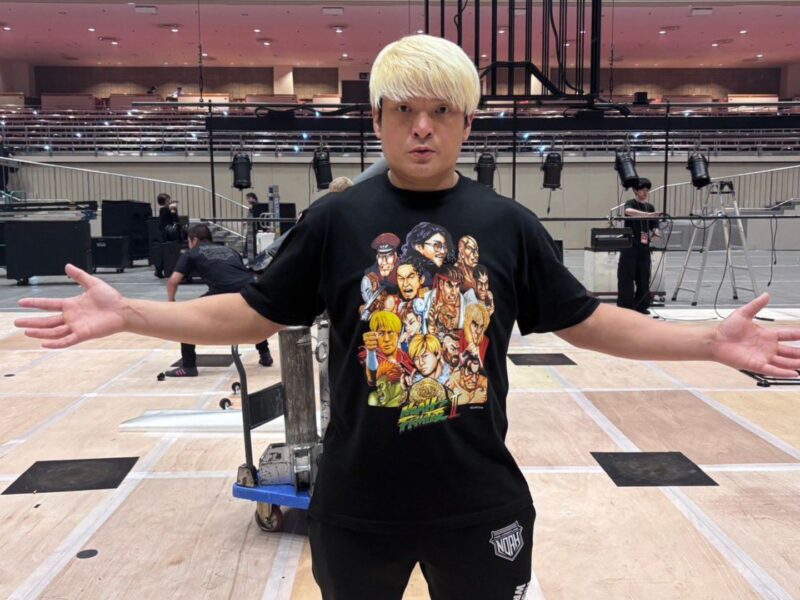 Man with platinum blond hair stands center stage with arms outstretched in a backstage rehearsal space; crew and equipment in the background.