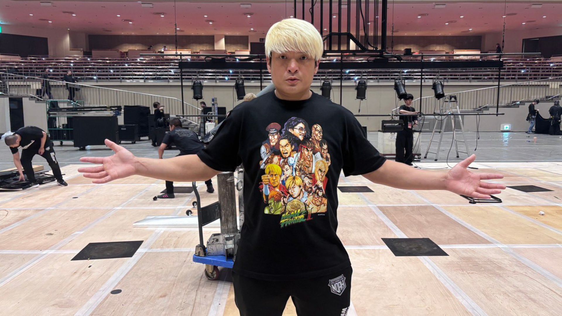 Man with platinum blond hair stands center stage with arms outstretched in a backstage rehearsal space; crew and equipment in the background.