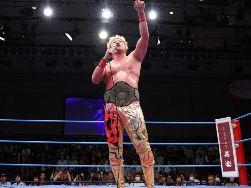 Wrestler stands in the ring with a championship belt around his waist, one arm raised in victory under bright arena lights.