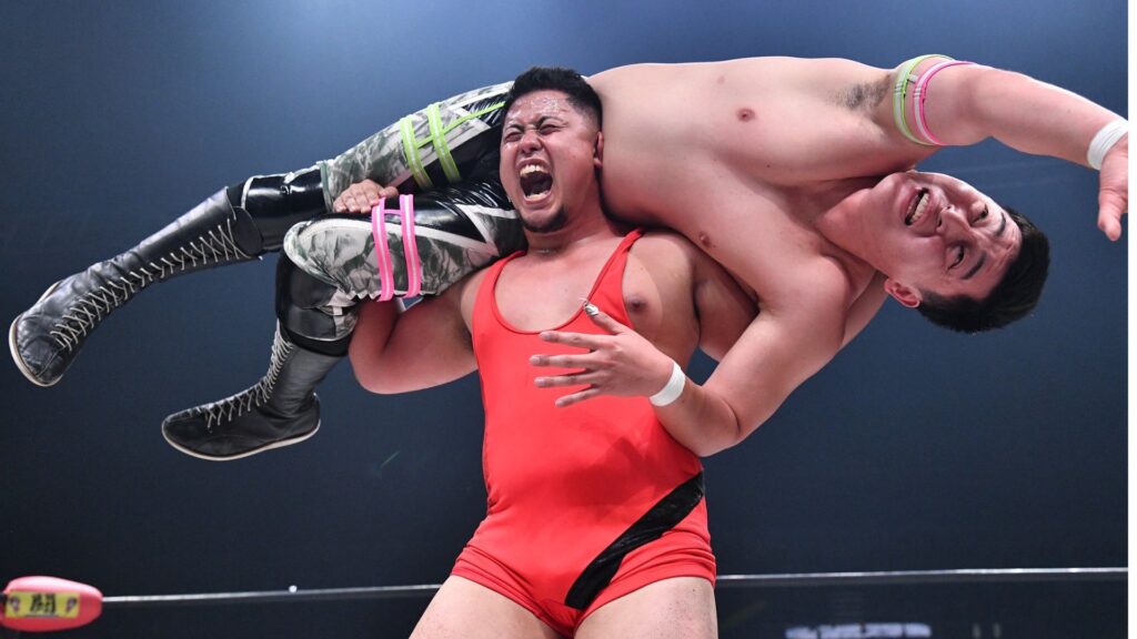 Two male wrestlers collide in a wrestling match: one in a red singlet lifts the other across his shoulders, the airborne wrestler grimacing in pain with camouflage pants and tall black boots gripping his opponent's arm.