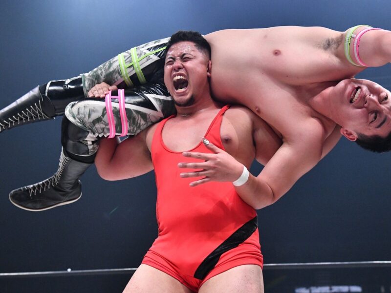 Two male wrestlers collide in a wrestling match: one in a red singlet lifts the other across his shoulders, the airborne wrestler grimacing in pain with camouflage pants and tall black boots gripping his opponent's arm.