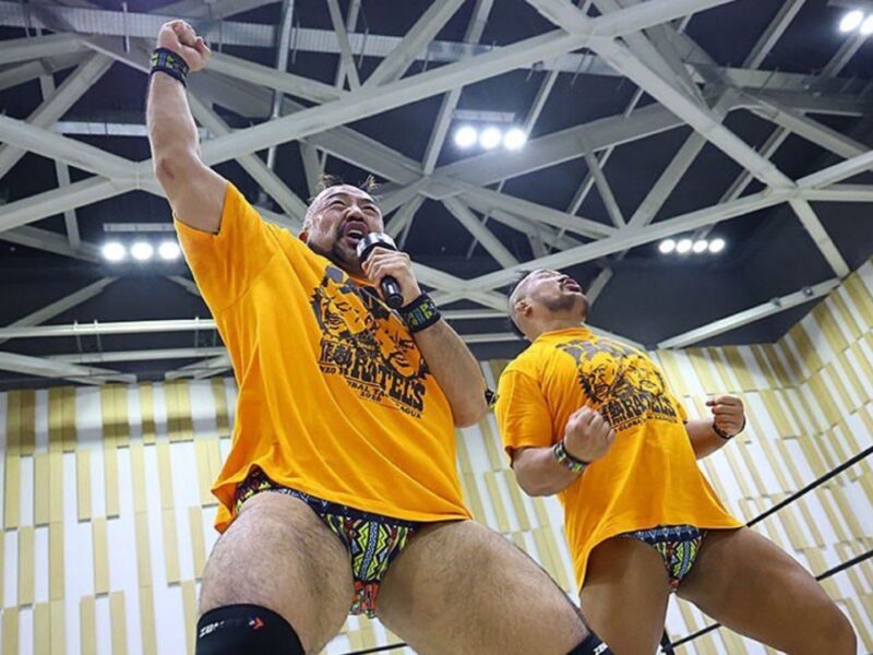 Two muscular wrestlers in matching orange shirts celebrate in a wrestling ring; one holds a microphone and raises a clenched fist.