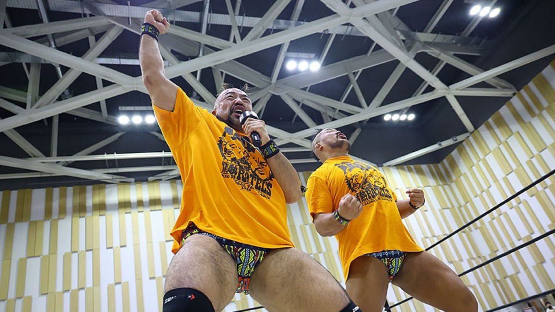 Two muscular wrestlers in matching orange shirts celebrate in a wrestling ring; one holds a microphone and raises a clenched fist.