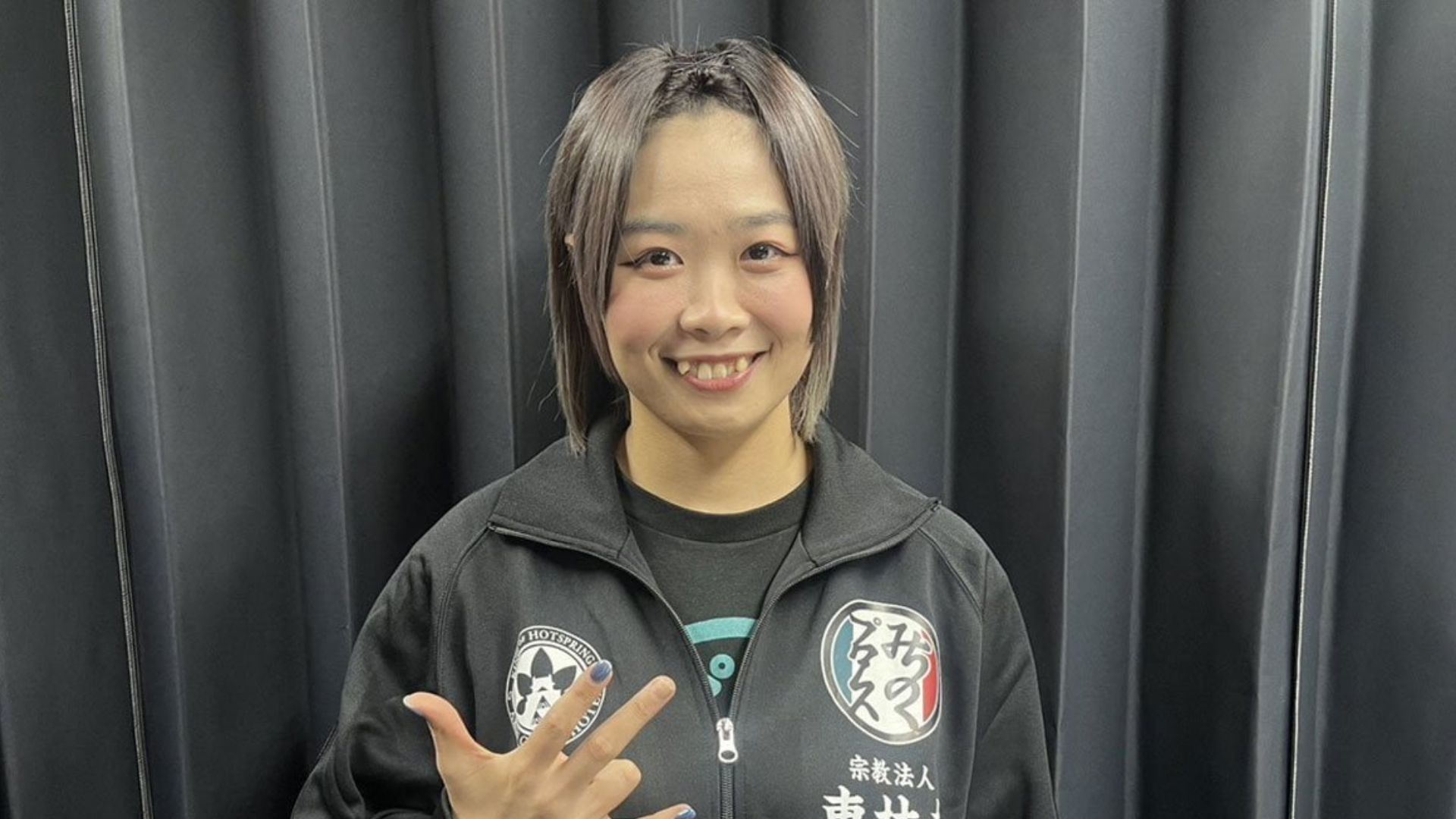 Smiling young woman in a black zip-up jacket with patches, making a peace sign at the camera against a dark curtain backdrop.