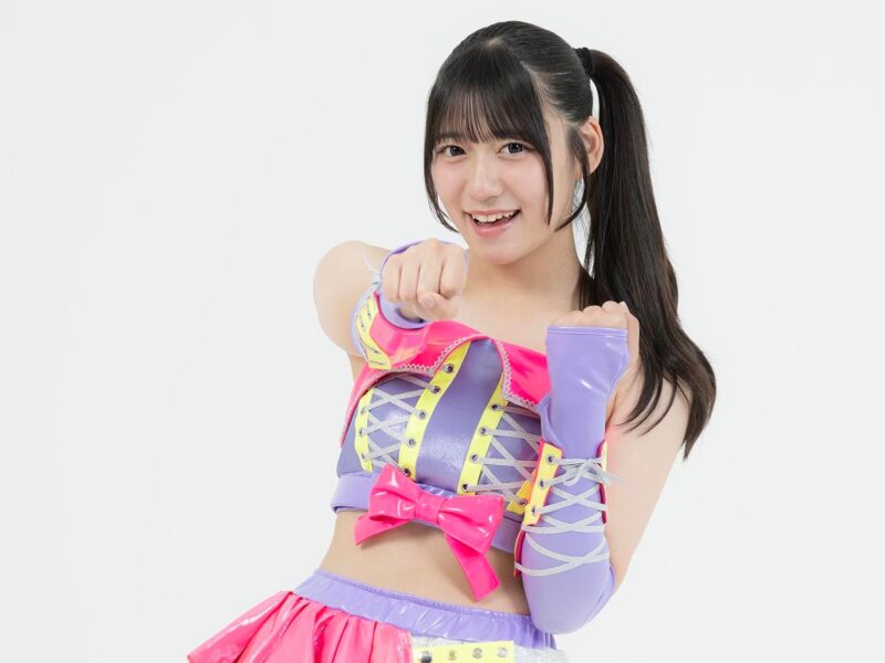 Young woman in a bright pink and lavender costume poses with fists up, smiling at the camera against a white background.