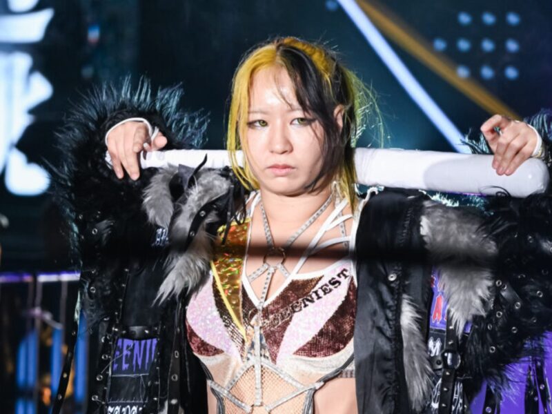 Female wrestler on stage with a black fur-trim jacket and a metallic pink-gold outfit, holding a white baton across her shoulders with a serious expression.