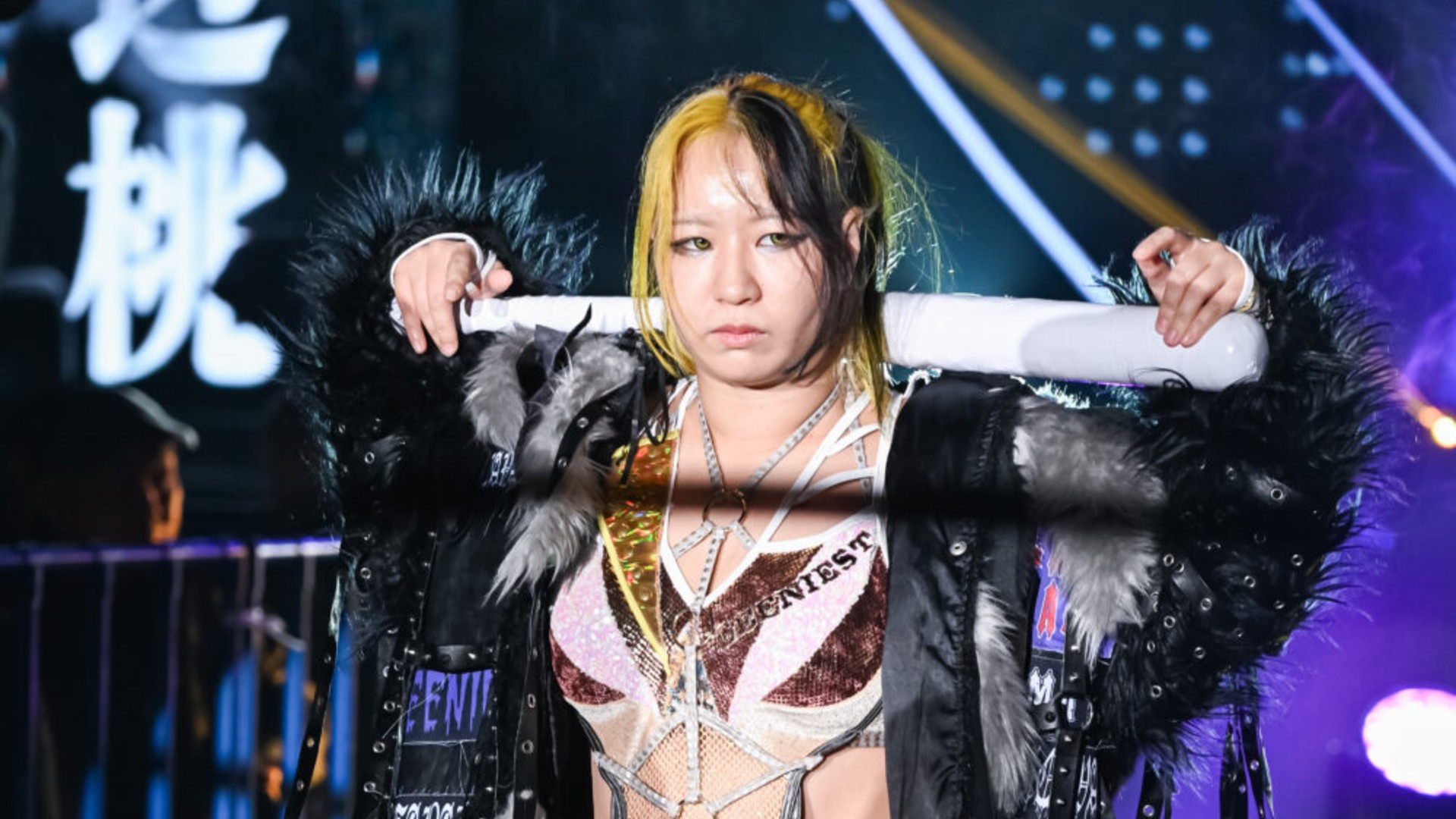 Female wrestler on stage with a black fur-trim jacket and a metallic pink-gold outfit, holding a white baton across her shoulders with a serious expression.