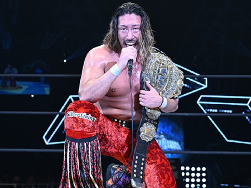 Wrestler in red trunks with long hair, holding a microphone and a championship belt in the ring.
