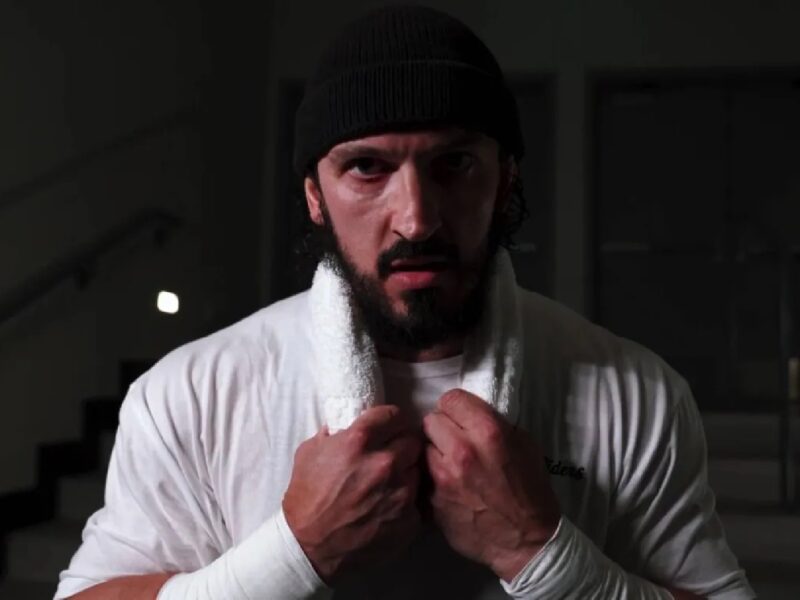 Man wearing a black beanie and white robe, gripping a towel at his chest in a dimly lit room.