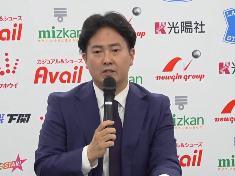 Man in a dark blue suit speaks into a handheld microphone at a press conference with sponsor logos on the backdrop behind him.