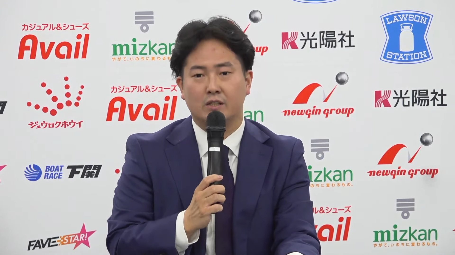 Man in a dark blue suit speaks into a handheld microphone at a press conference with sponsor logos on the backdrop behind him.