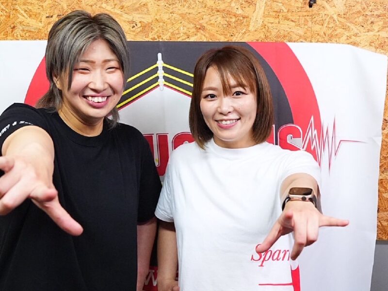 Two women pose for a selfie, smiling at the camera as they extend their arms toward the lens, with a white-and-red banner and wood-panel backdrop behind them.