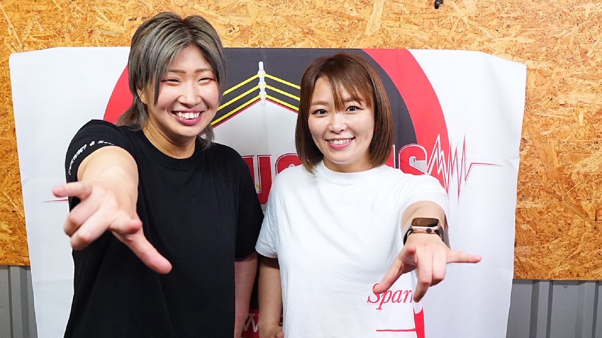 Two women pose for a selfie, smiling at the camera as they extend their arms toward the lens, with a white-and-red banner and wood-panel backdrop behind them.