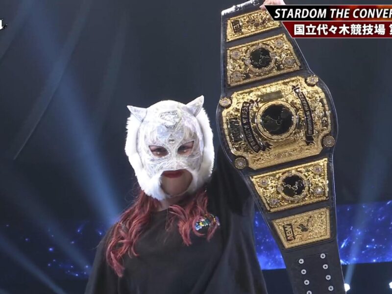 Masked female wrestler raises a gold championship belt on stage at Stardom The Conversion 2026.