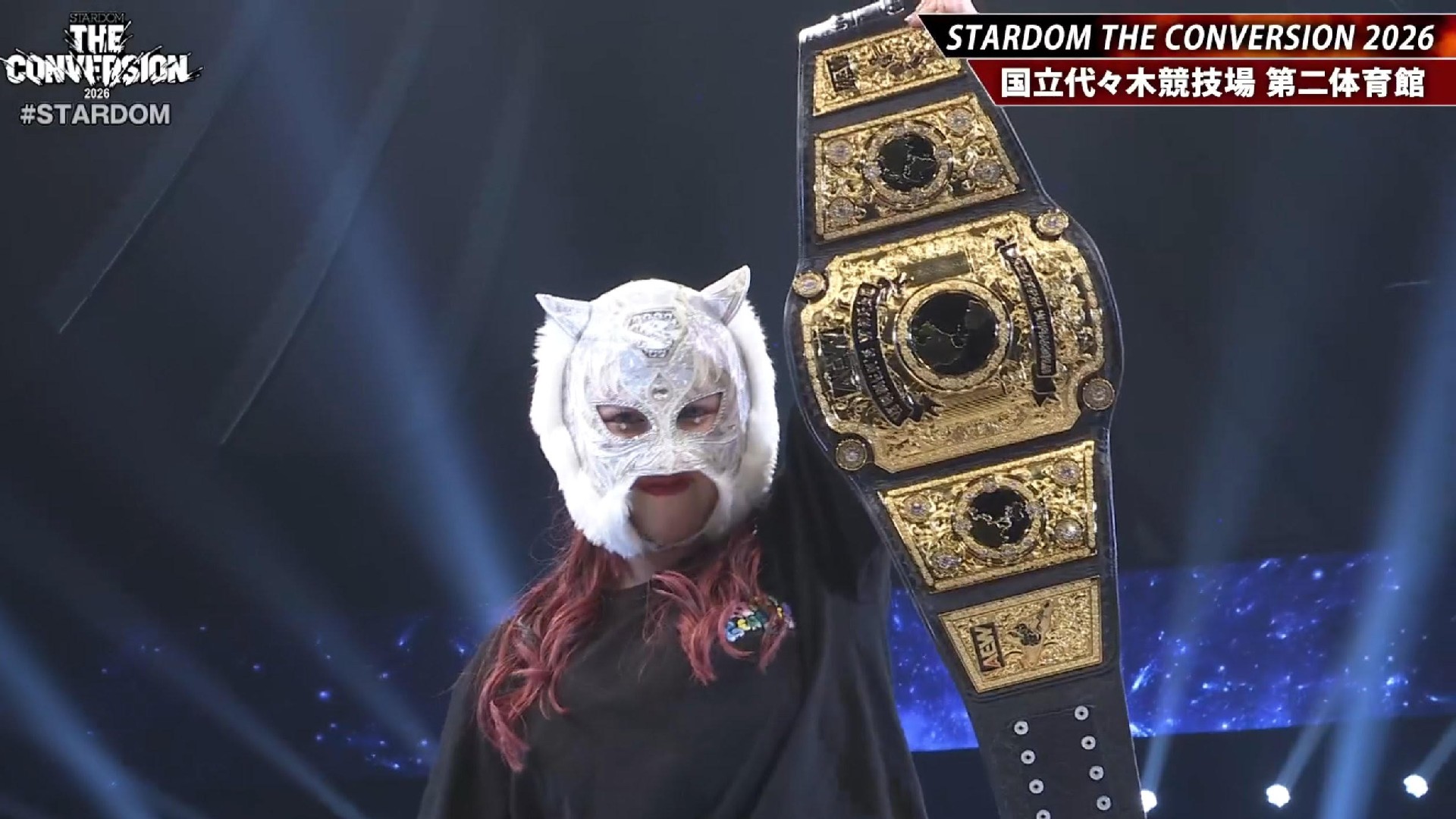 Masked female wrestler raises a gold championship belt on stage at Stardom The Conversion 2026.