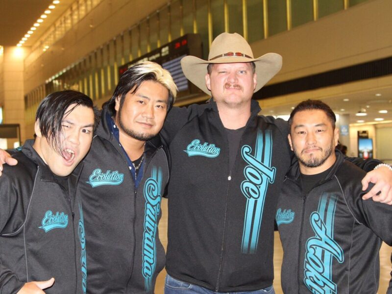 Four men in matching black teal Evolution jackets pose with arms around each other in an airport terminal, smiling for the photo.