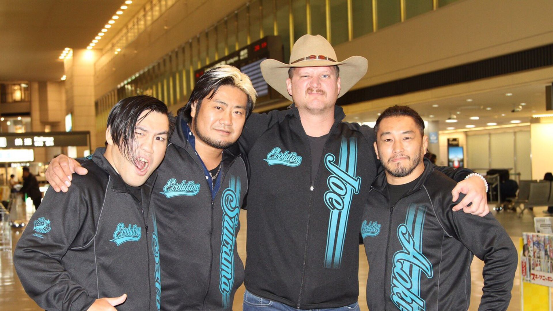 Four men in matching black teal Evolution jackets pose with arms around each other in an airport terminal, smiling for the photo.