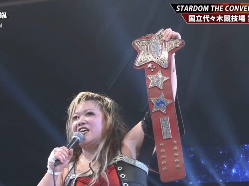 Female wrestler on stage, microphone in hand, raising a large, ornate championship belt in victory.
