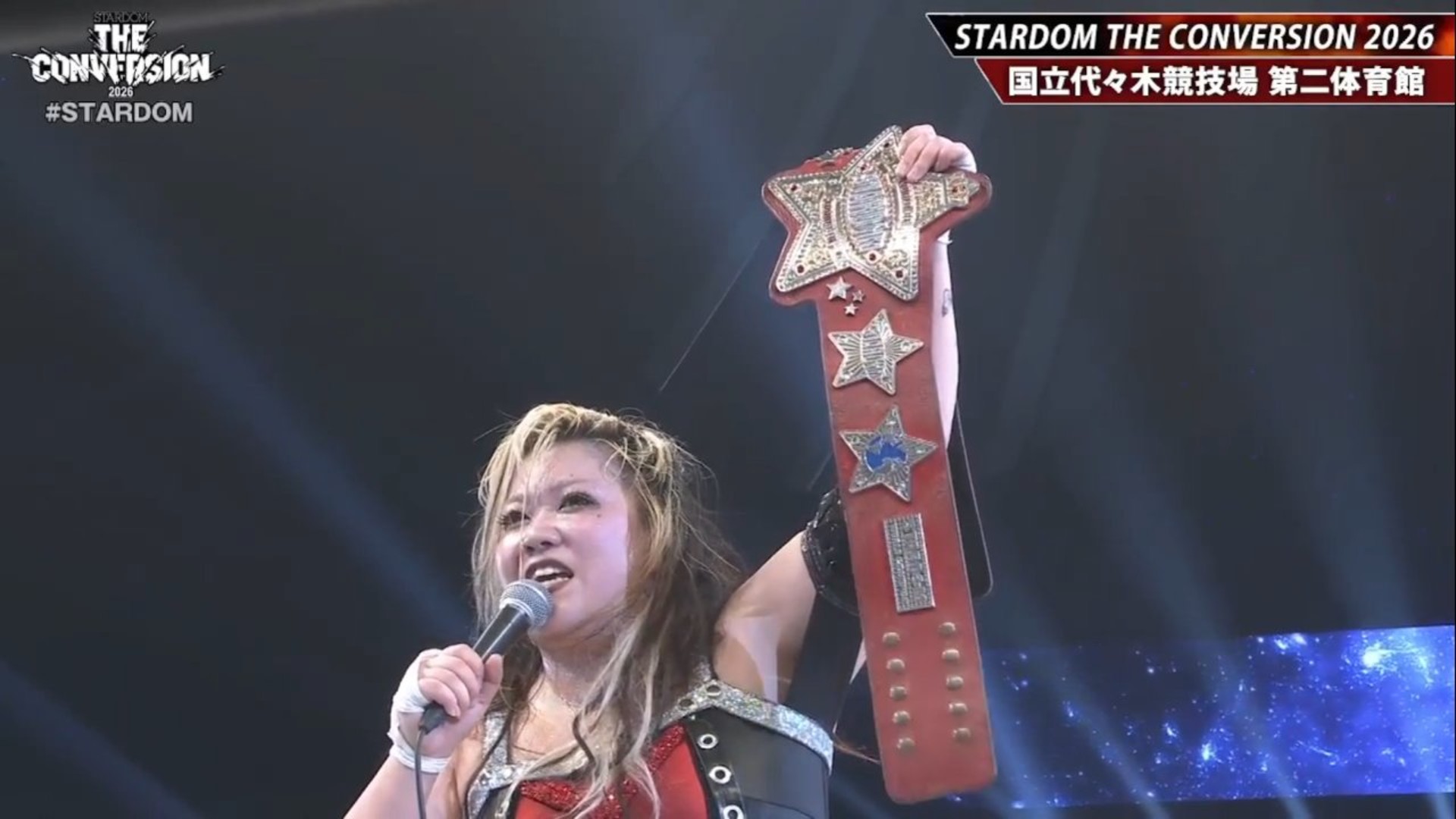 Female wrestler on stage, microphone in hand, raising a large, ornate championship belt in victory.