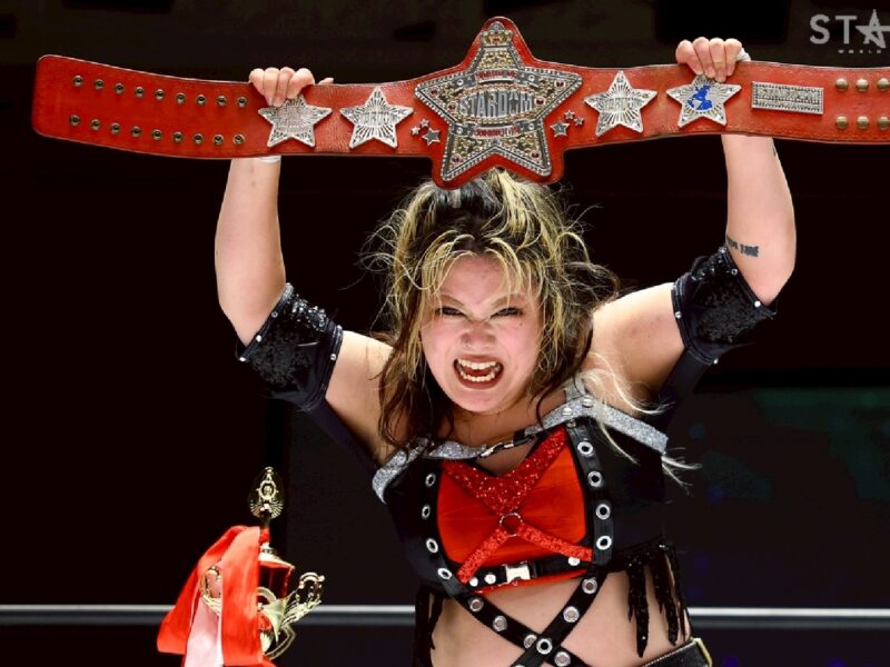 Female professional wrestler raises a large red championship belt overhead in victory.