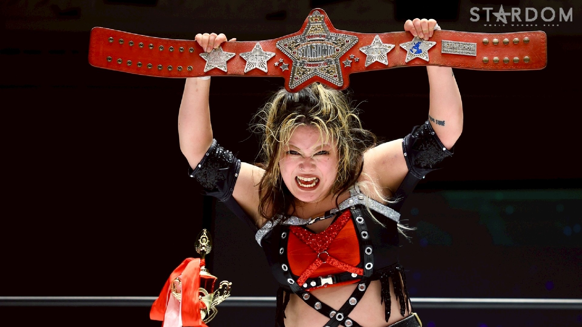 Female professional wrestler raises a large red championship belt overhead in victory.