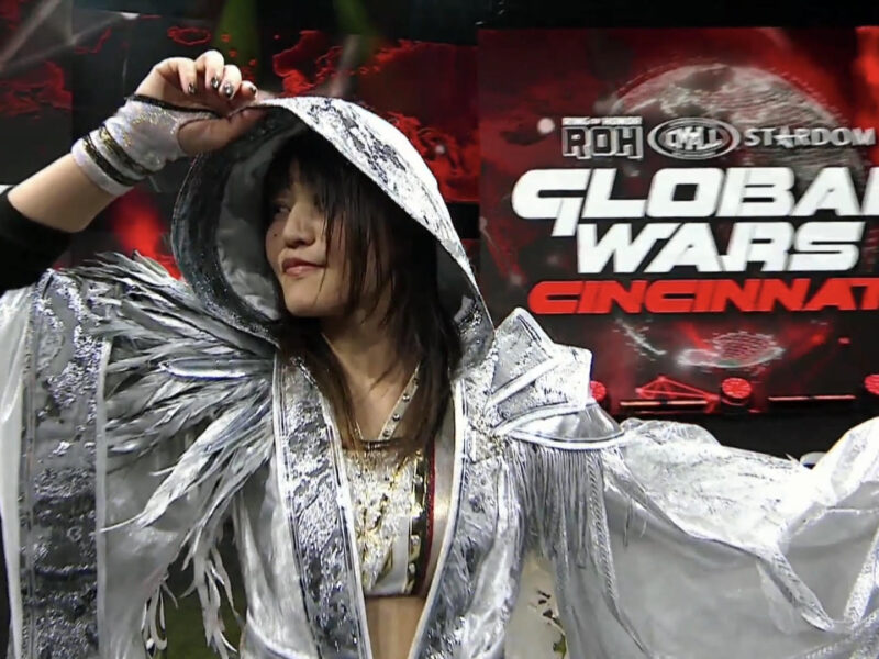 Wrestler in a shiny silver hooded costume raises an arm on stage with a 'Global Wars Cincinnati' backdrop behind.