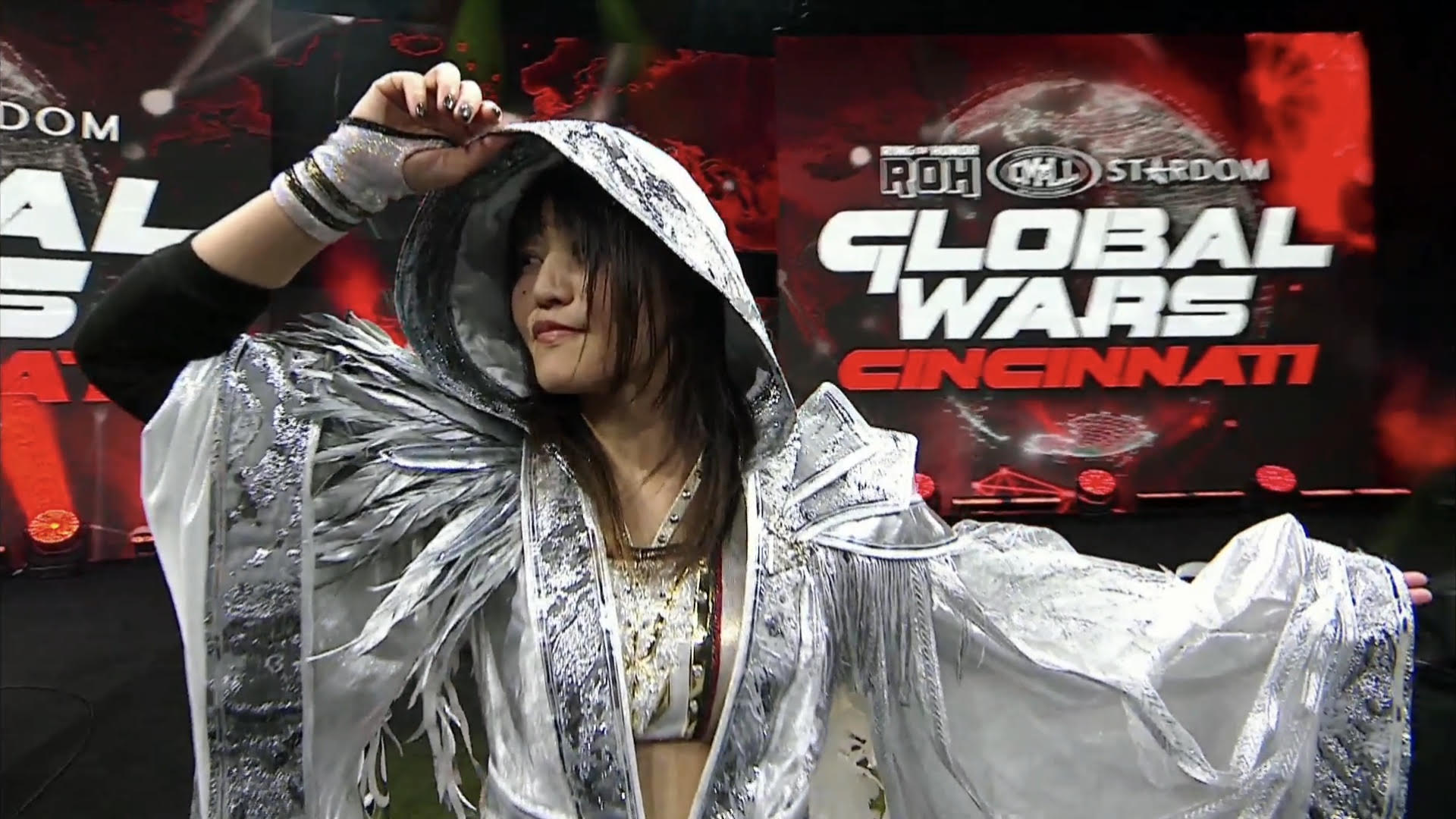 Wrestler in a shiny silver hooded costume raises an arm on stage with a 'Global Wars Cincinnati' backdrop behind.