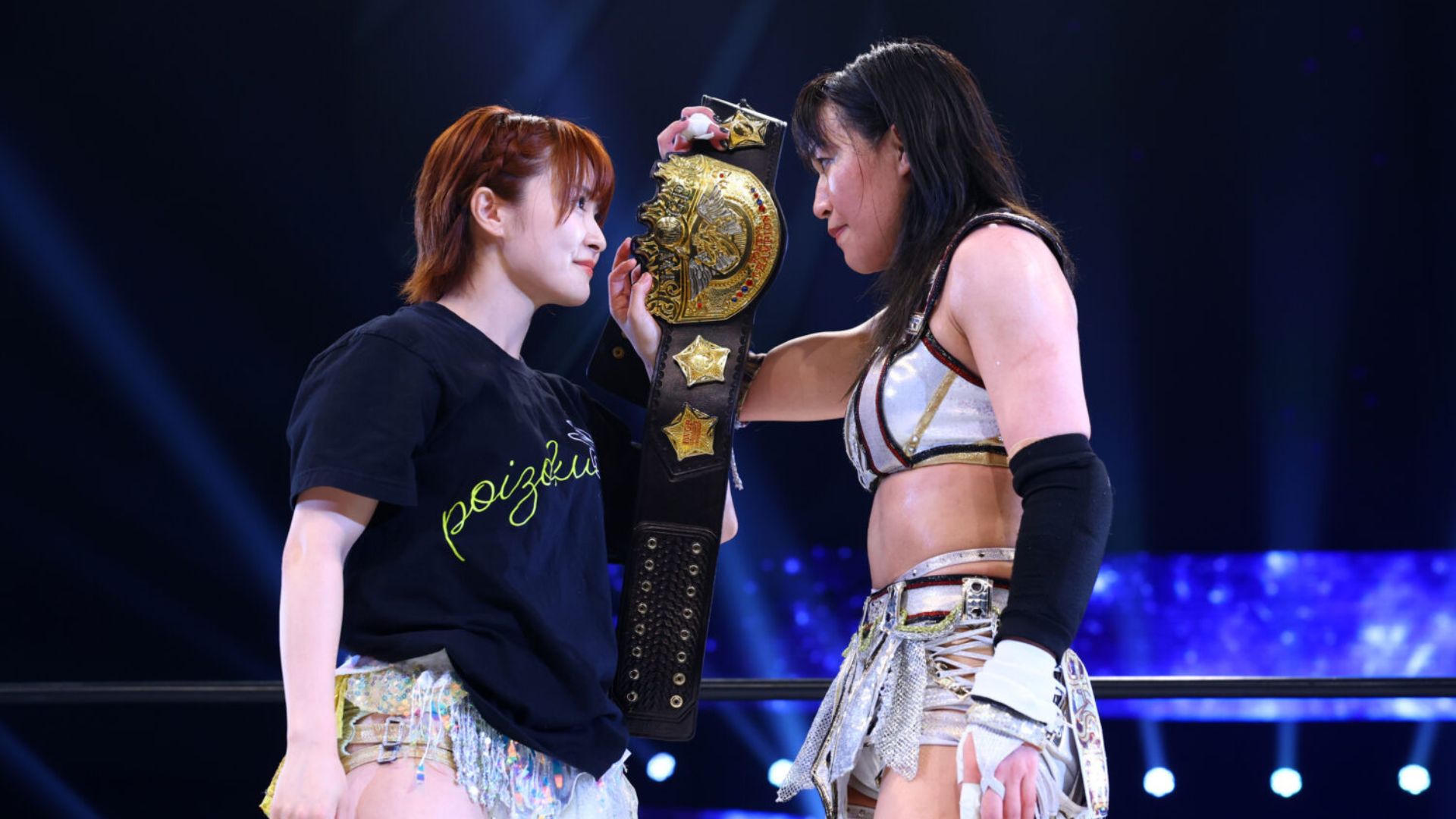 Two female wrestlers face off in the ring, with a gold championship belt held between them.