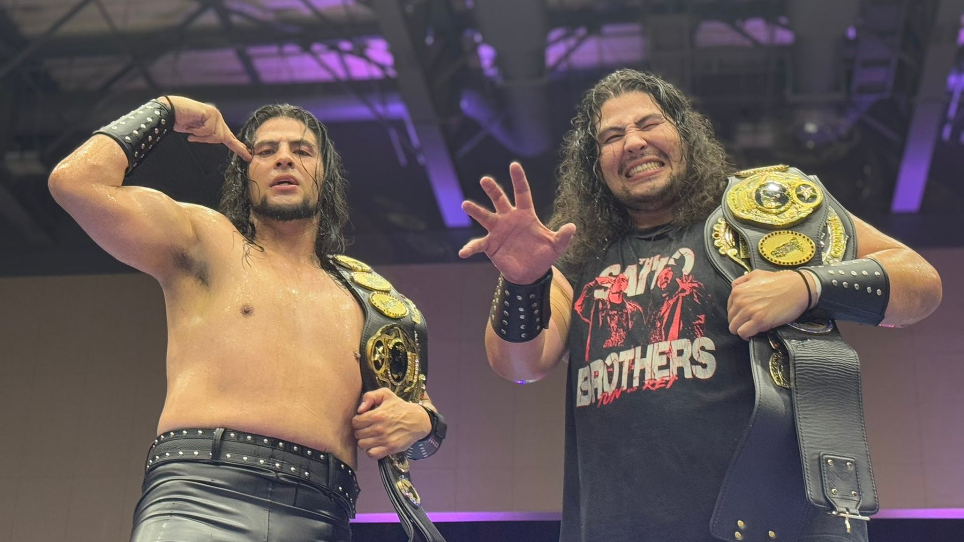 Two wrestlers celebrate with championship belts, one shirtless in studded pants and the other in a black Brothers shirt, both showing triumphant poses under arena lights.