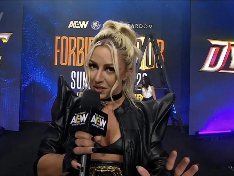 Female wrestler wearing a black leather jacket speaks to the camera, holding an AEW microphone on a blue stage backdrop.